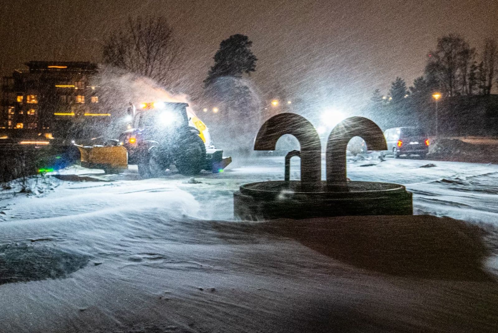 Traktor med brøyteskjær rydder snø i snøfokk en vinterkveld, med boligblokker i bakgrunnen