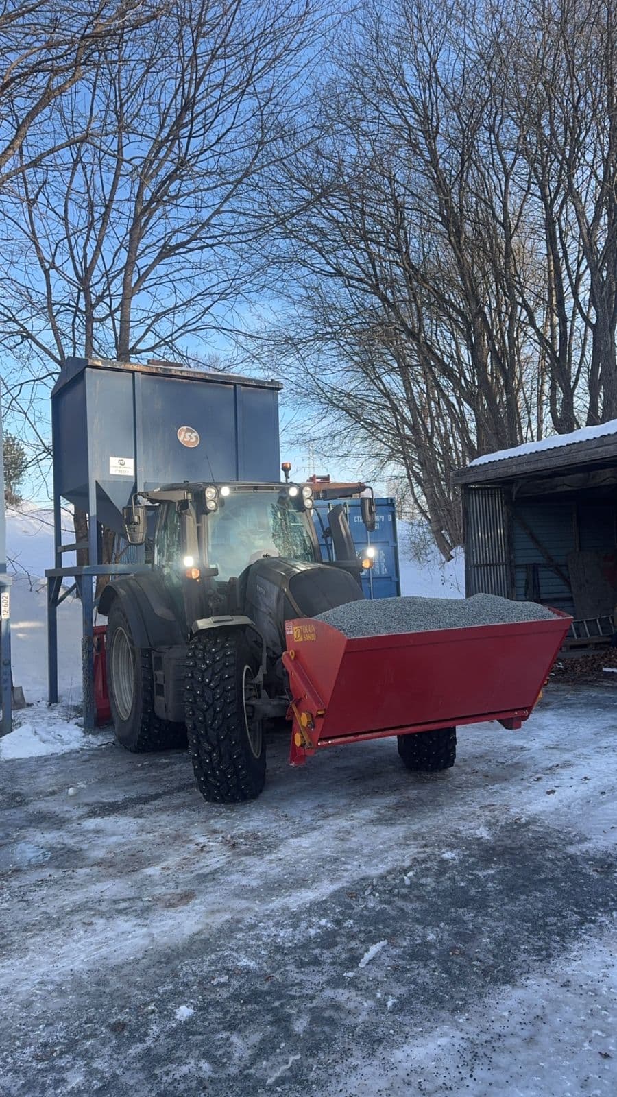 Traktor med strøkasse full av grus foran en strøsilo