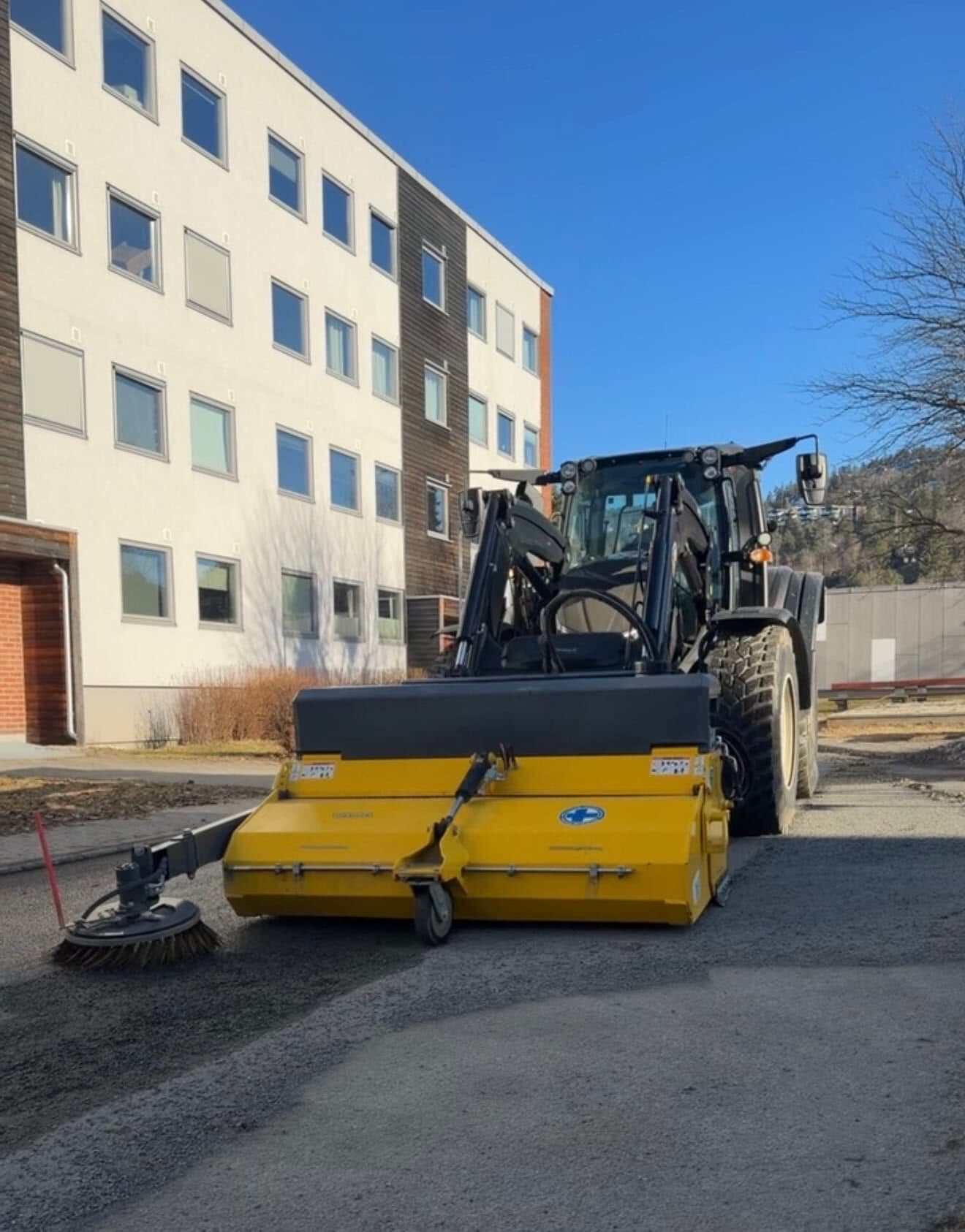 Traktor med feiemaskin feier bort strøsand ved en boligblokk om våren